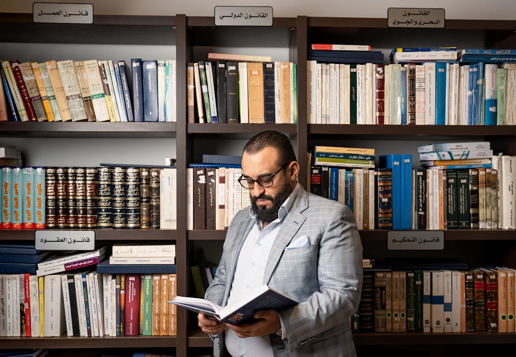 A bearded businessman in a suit reads a book in a library with shelves full of books.