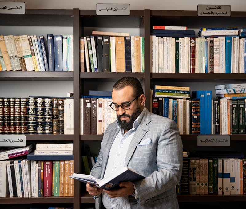 A bearded businessman in a suit reads a book in a library with shelves full of books.