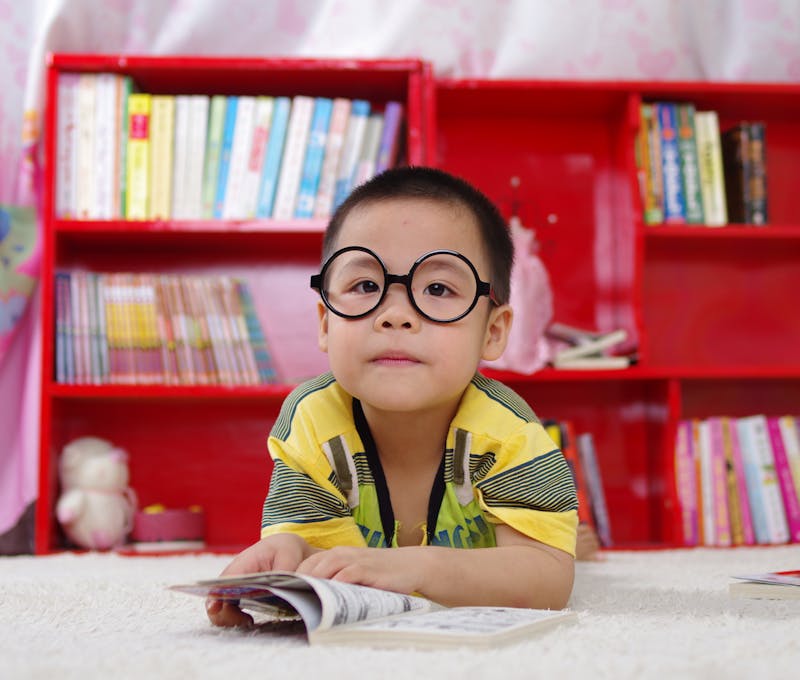 A cute child in glasses reading a book, surrounded by colorful shelves. Perfect shot of childhood learning indoors.