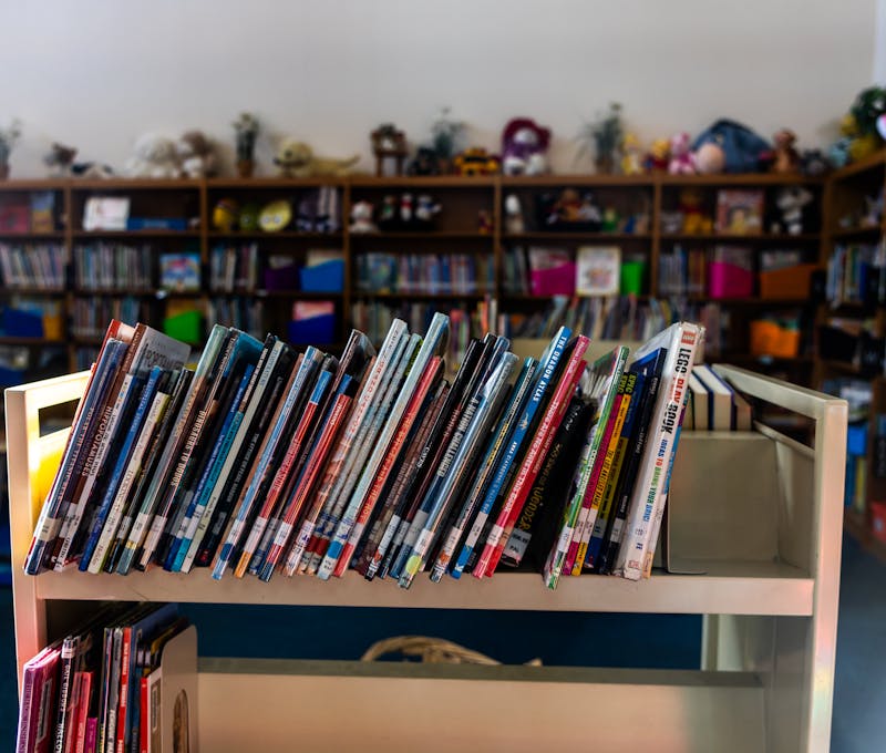 A warm library interior featuring a book cart in focus with colorful children's books on shelves.