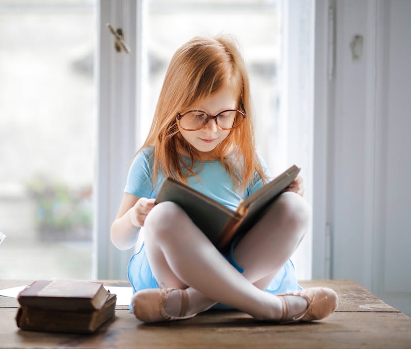 Adorable young girl wearing glasses, reading a book indoors by a window. Captures innocence and focus.
