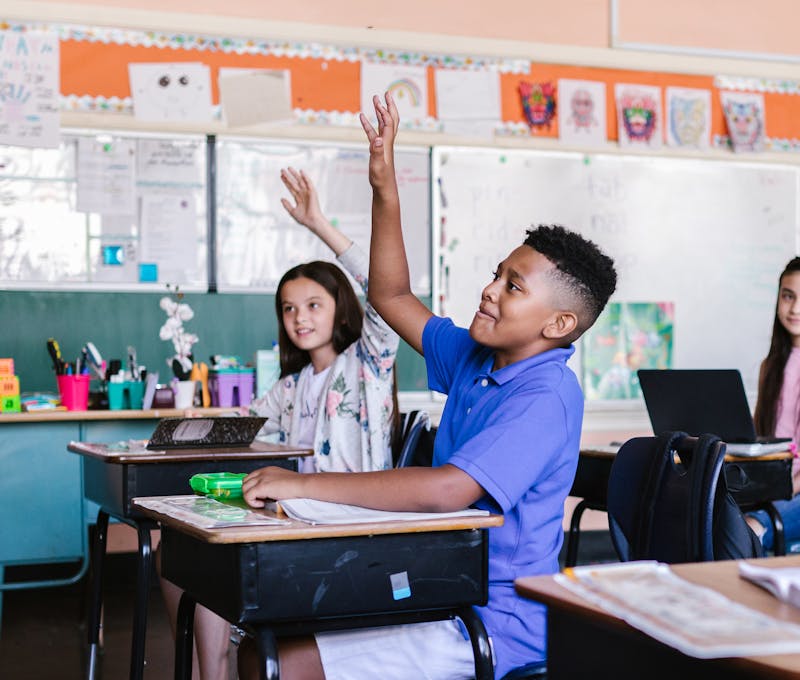 Children in a lively classroom raising hands during an interactive lesson.