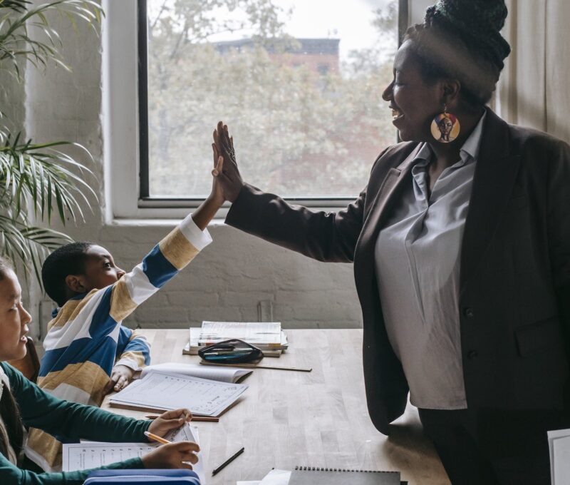 Side view of smiling black boy giving high five to teacher while sitting with diverse classmate girl at desk in classroom