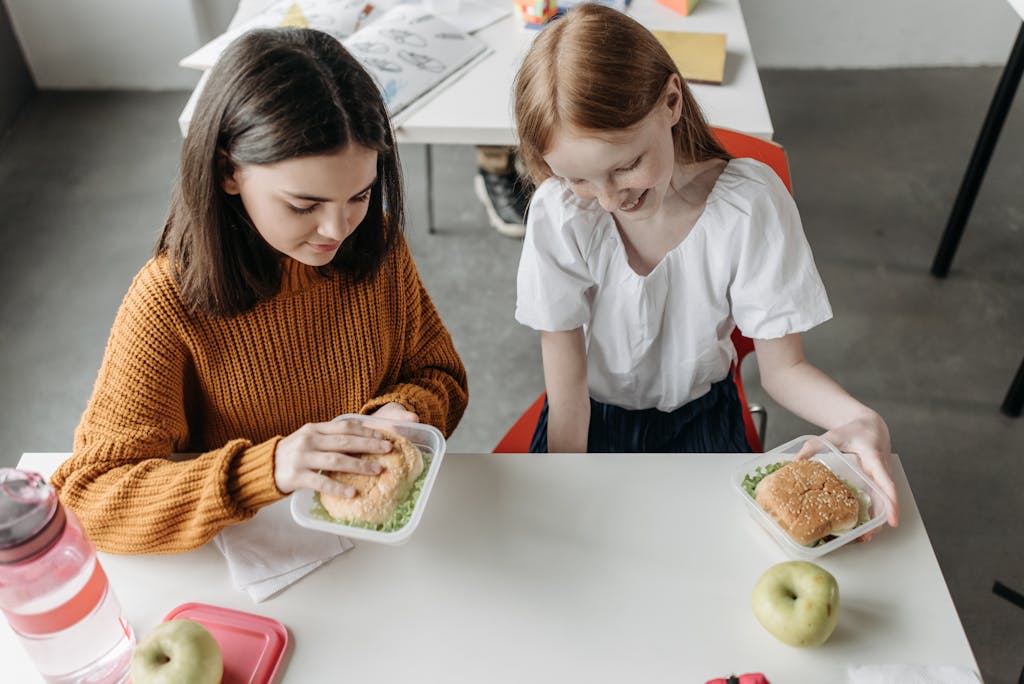 Two young girls enjoying lunch together at a school desk with sandwiches and apples.