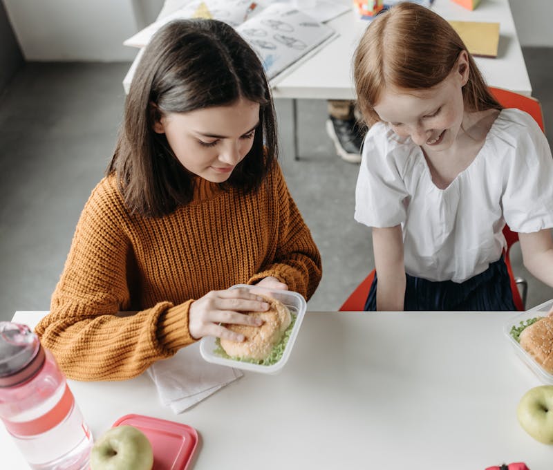 Two young girls enjoying lunch together at a school desk with sandwiches and apples.