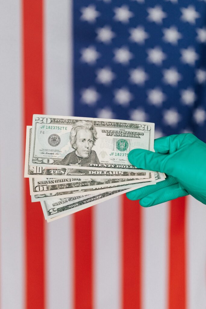 Unrecognizable person in medical disposable gloves demonstrating paper banknotes against multicolored American flag on background