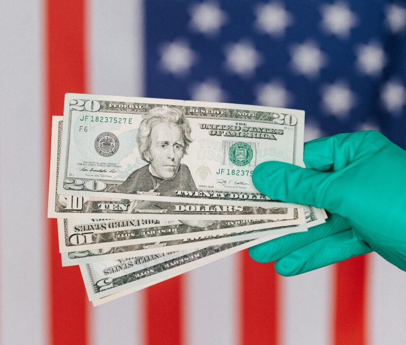 Unrecognizable person in medical disposable gloves demonstrating paper banknotes against multicolored American flag on background