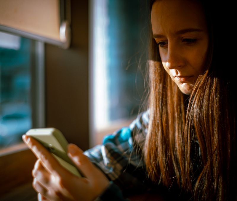 Young woman looking at smartphone with sunlight streaming in through a window.